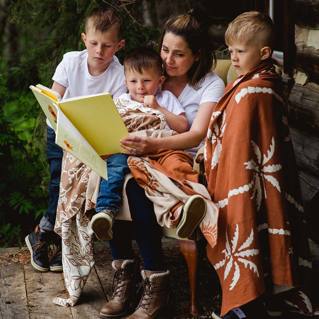 Young mum reading her small children a book sitting in an armchair surrounded by her children who are draped in Snuggly Jacks Organic Knitted Blankets