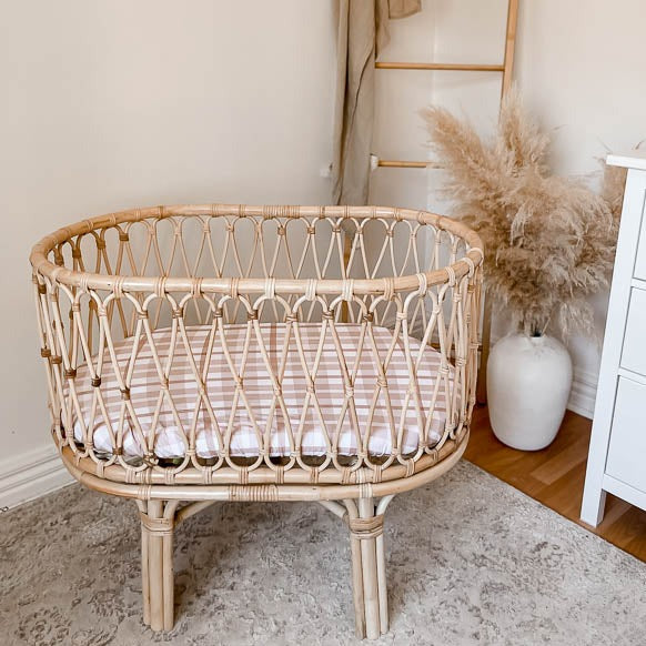 Modern Boho nursery with a rattan bassinet, decorative bamboo ladder in the back ground, a vase with decorative grass and a  white chest of draws on the very right side of the space.