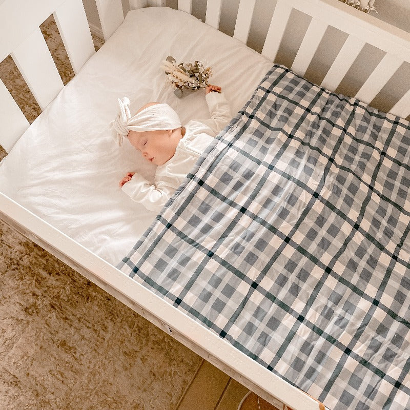 Baby wearing a white top knot laying in a white cot snug under a blue plaid crib quilt.