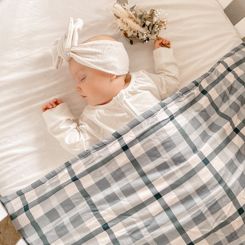 Baby wearing a white top knot sleeping under a blue plaid cotton crib quilt