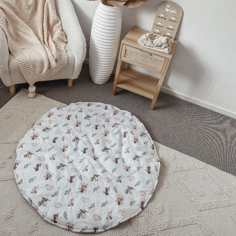 A 100% cotton playmat with adorable dragons laid out on the floor of a modern nursery with and arm chair in one corner and a pine draw with rattan details
