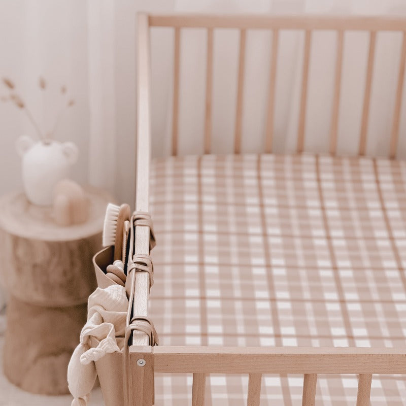 Close up of a wooden cot with a nursery organiser hanging on one side a wooden bedside table in the back ground and a brown and white plaid fitted crib sheet