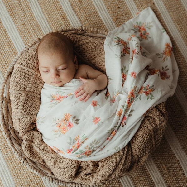 Snuggly Jacks Bloom Floral Muslin Wrap tightly wrapping a little baby sleeping on top of brown knitted blanket in a moses basket.